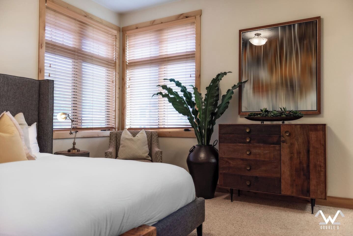 Quarto com cama, mesa de cabeceira, luminária, cômoda e uma janela grande com persianas.
