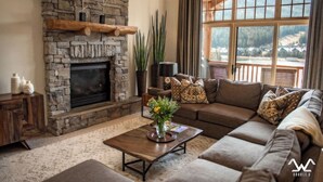 A cozy living room with a stone fireplace, a large window with a view of a mountainous landscape, and a comfortable brown sofa with patterned cushions. - Luxurious King en Suites in a Pristine Penthouse! (Big Sky)