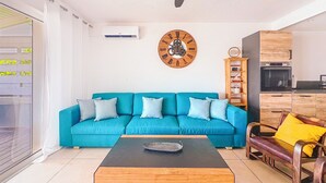 A living room with a teal sofa, a wooden coffee table, a wooden side table, a wooden chair with a yellow cushion, a wooden clock, and a kitchen area with wooden cabinets and appliances. - Villa Little Paradise - BIG DISCOUNT MARCH - APRIL !! (Collectivity of Saint Martin)