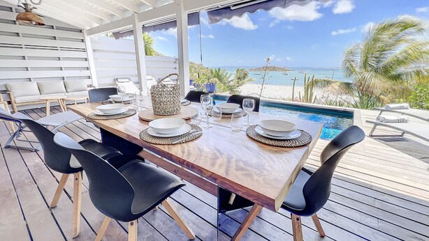 An outdoor dining area with a wooden table set for four, surrounded by black chairs, overlooking a pool and beach. - Villa Little Paradise - BIG DISCOUNT MARCH - APRIL !! (Collectivity of Saint Martin)