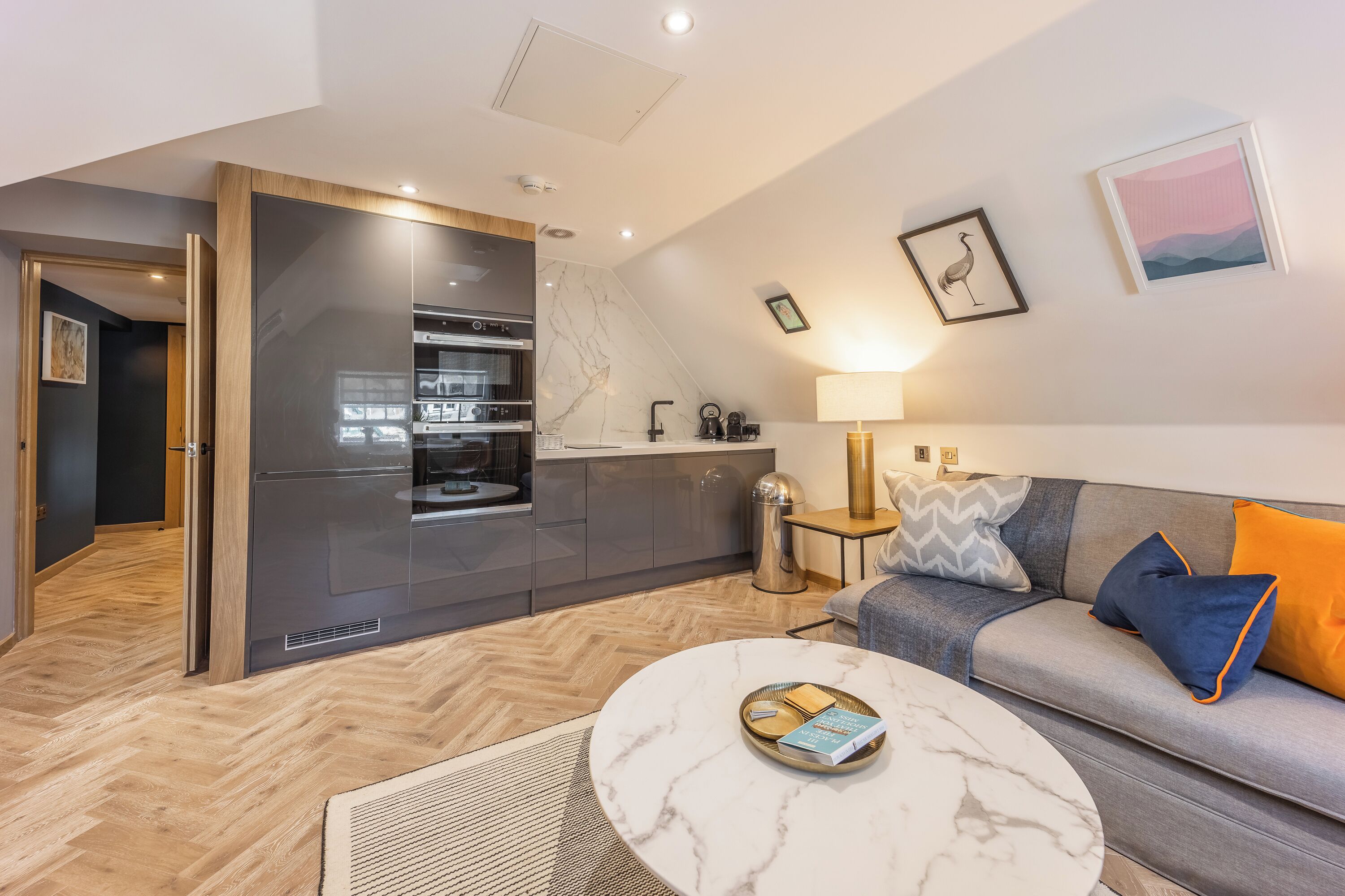 A modern kitchen with built-in appliances, a grey sofa with colorful cushions, and a marble coffee table.