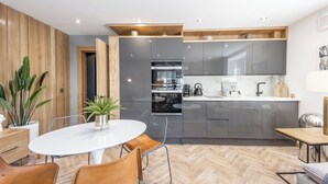 Une cuisine moderne avec des meubles gris, un four encastré et un plan de travail blanc. On y trouve un coin repas avec une table ronde et des chaises, ainsi qu'un plancher en bois.