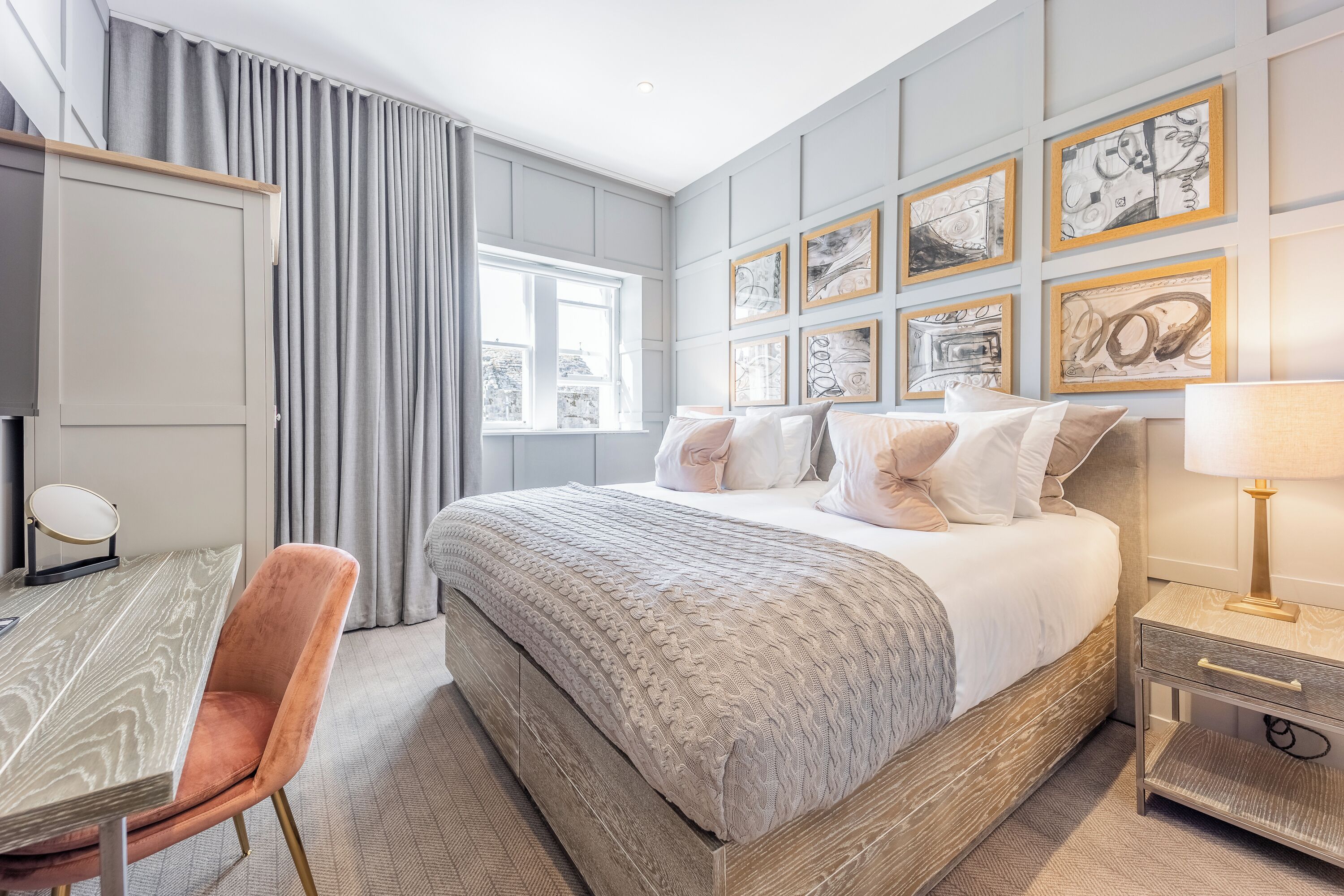 Una habitación de hotel moderna con una cama grande, un escritorio y una silla. Hay varios cuadros en la pared y una ventana con cortinas.