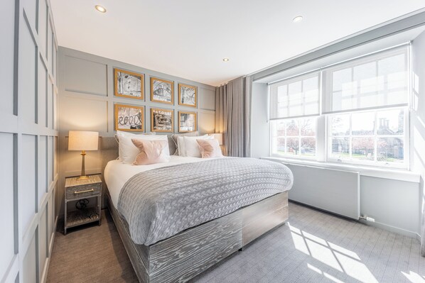 A modern bedroom with a large bed, a nightstand, a window with a view, and framed artwork on the wall.
