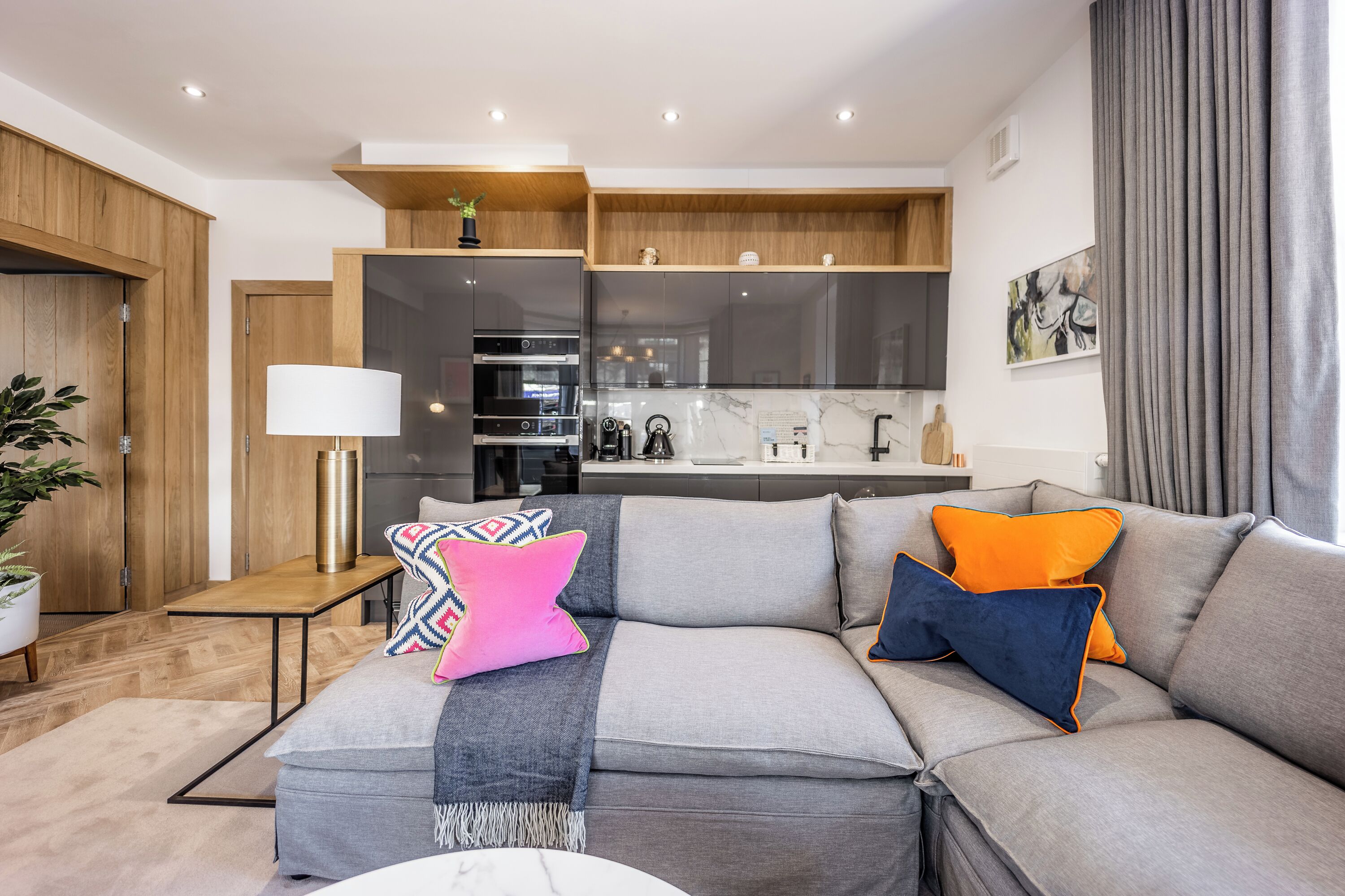 A modern living room with a grey sofa, colorful cushions, a small table with a lamp, and a kitchen area in the background.