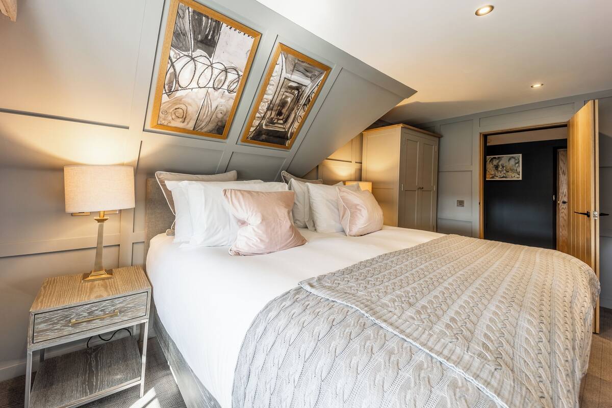 Ein ordentlich eingerichtetes Schlafzimmer mit einem großen Bett, einem Nachttisch und einem Beistelltisch. Über dem Bett hängen zwei gerahmte Kunstwerke, und eine Tür führt in einen anderen Raum.