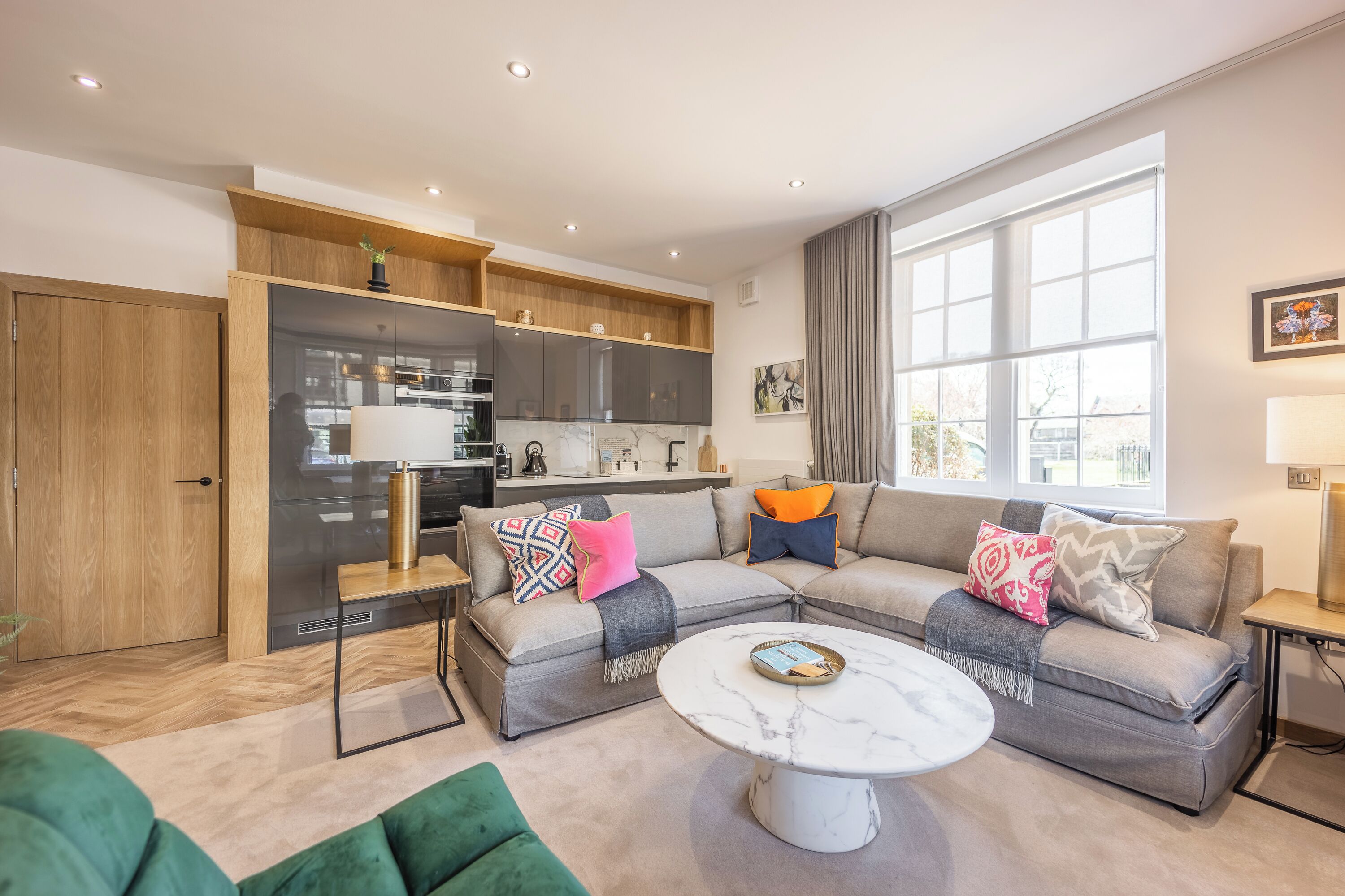 A modern living room with a sectional sofa, a marble coffee table, and a kitchen area with wooden cabinets and a built-in oven.