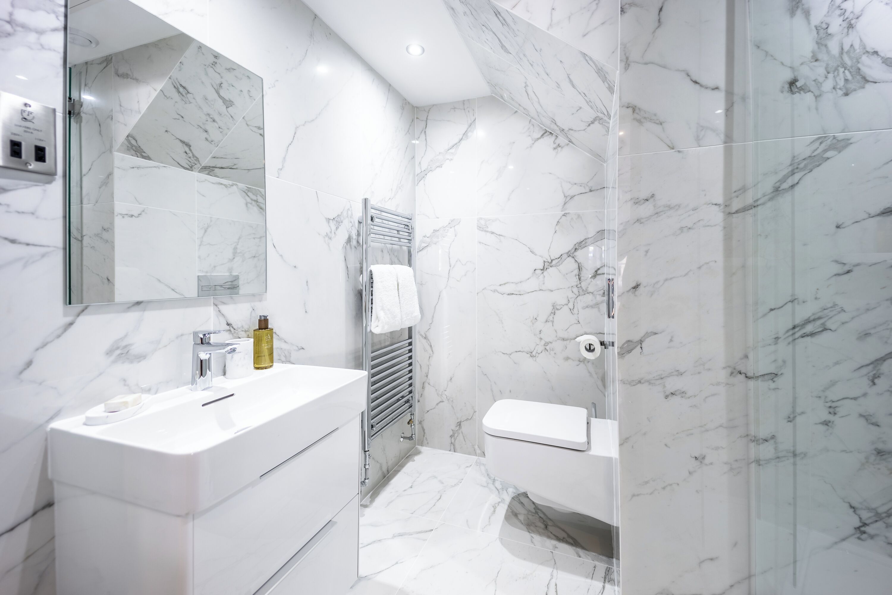 Un baño moderno con piso de mármol, un lavamanos blanco, un inodoro y una zona de ducha con mampara de vidrio.