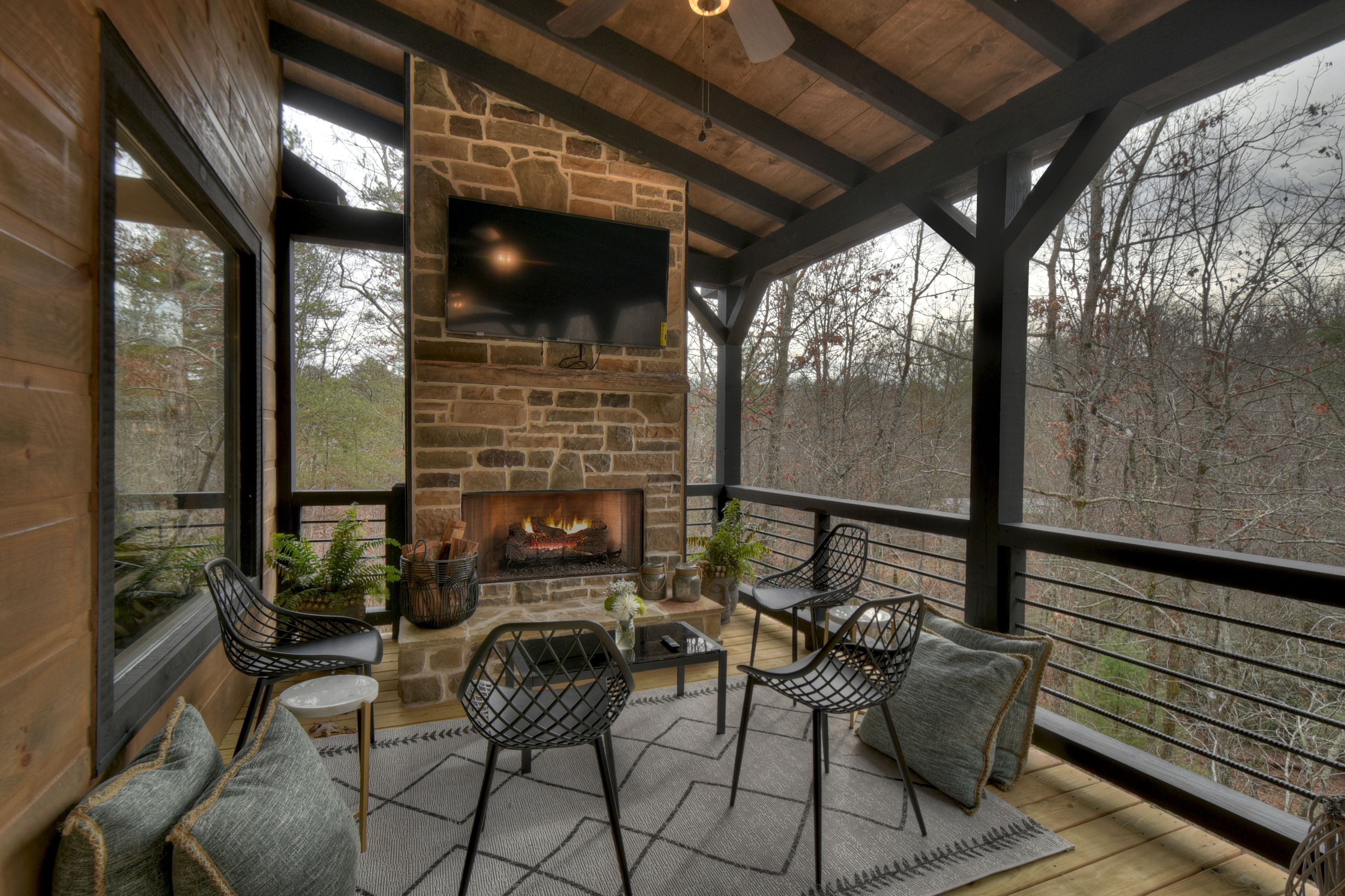 A covered outdoor seating area with a stone fireplace, black metal chairs, and a television mounted on the wall.