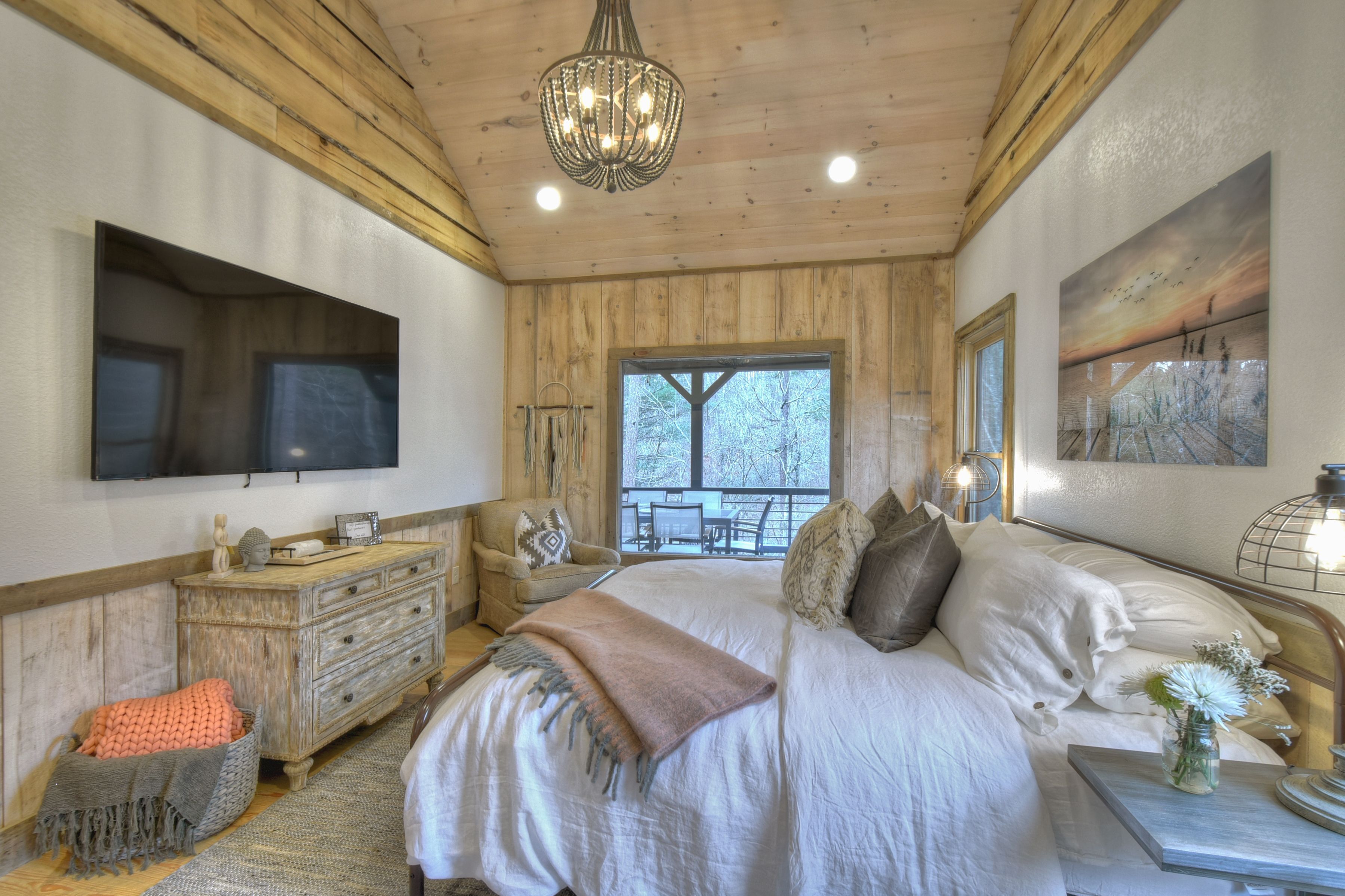 A bedroom with a large bed, wooden walls, a chandelier, a TV, and a window with a view of trees.