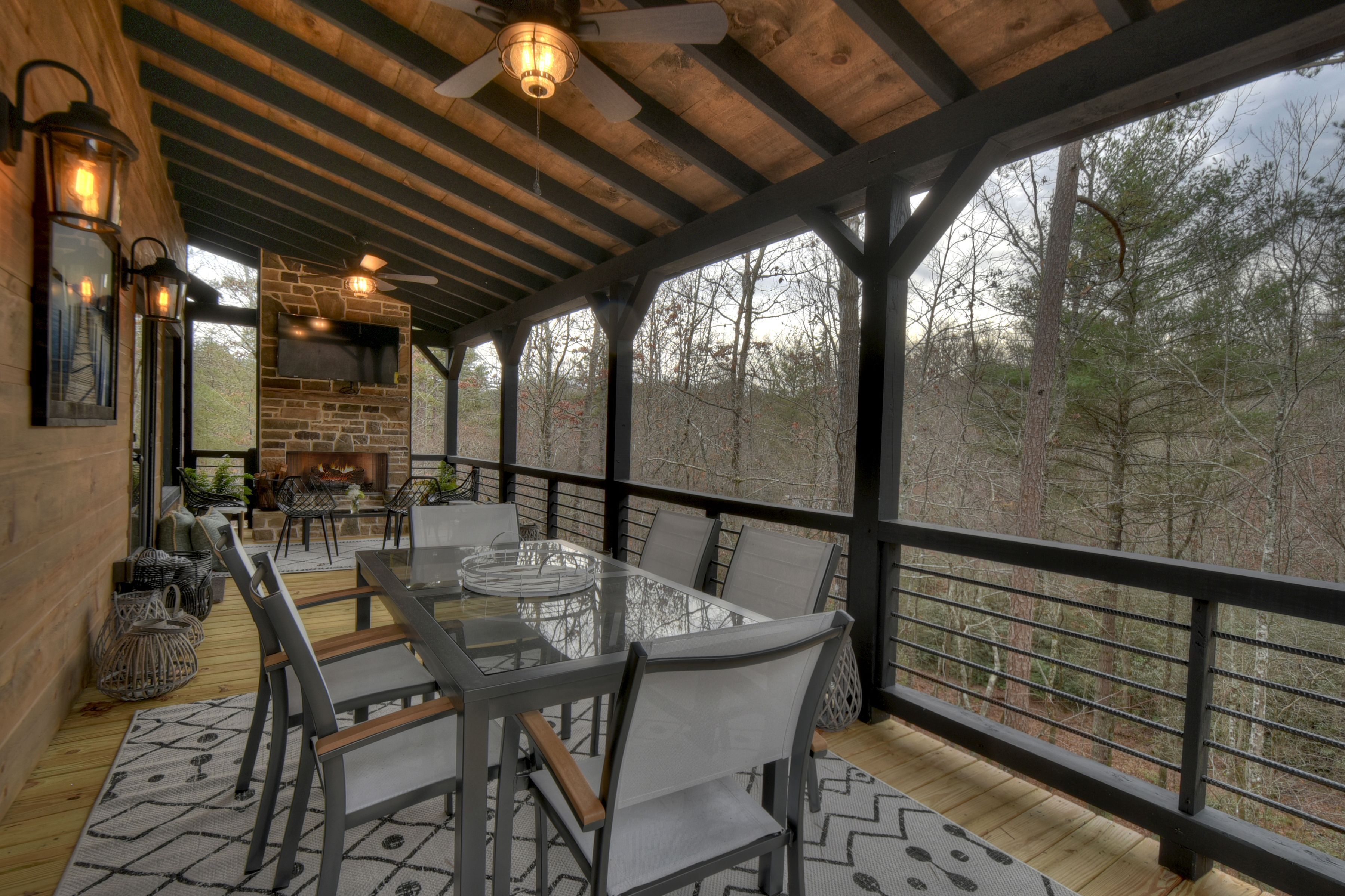 A covered outdoor dining area with a stone fireplace, wooden ceiling beams, and a view of trees.