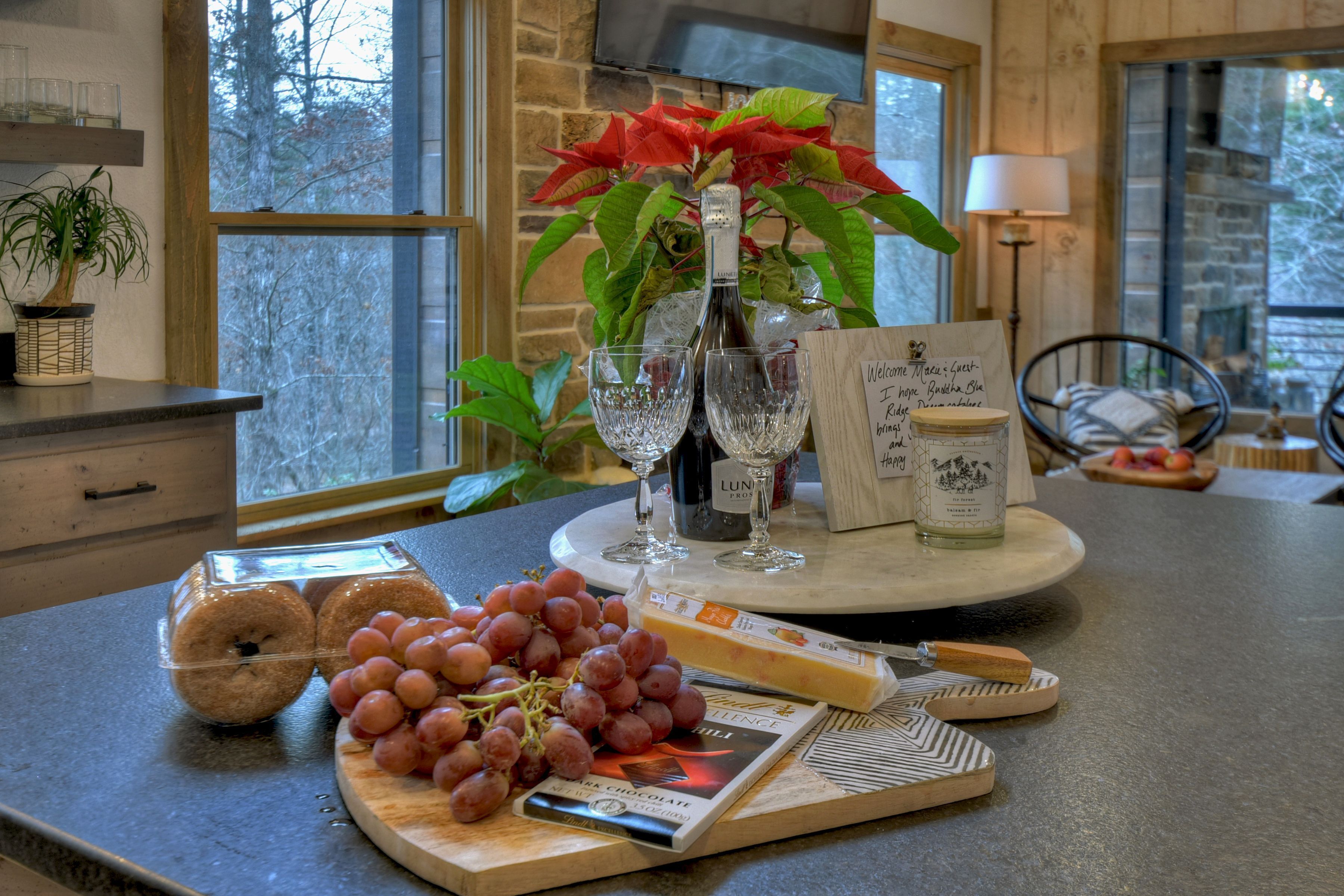 A kitchen counter with a bottle of wine, glasses, a cutting board with grapes and cheese, and a welcome sign.
