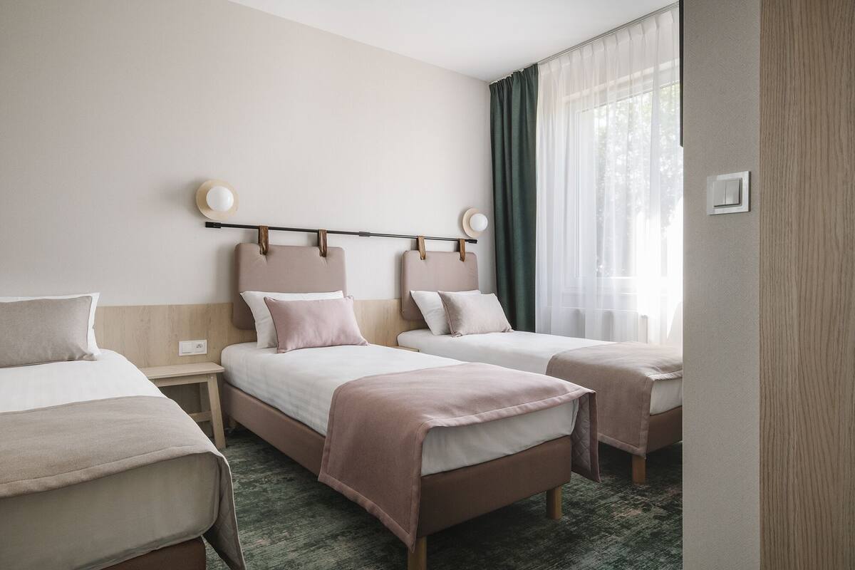Een hotelkamer met twee bedden, een groot raam met dunne gordijnen, een nachtkastje met een lamp en een wandlamp.