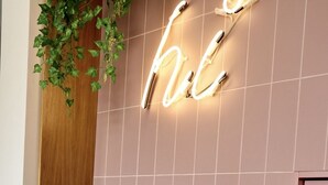 Una persona de pie frente a un letrero de neón que dice “love” sobre un muro de azulejos, con una planta en maceta a la izquierda.