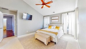 Un dormitorio con cama, ventilador de techo, televisor y ventana con cortinas.