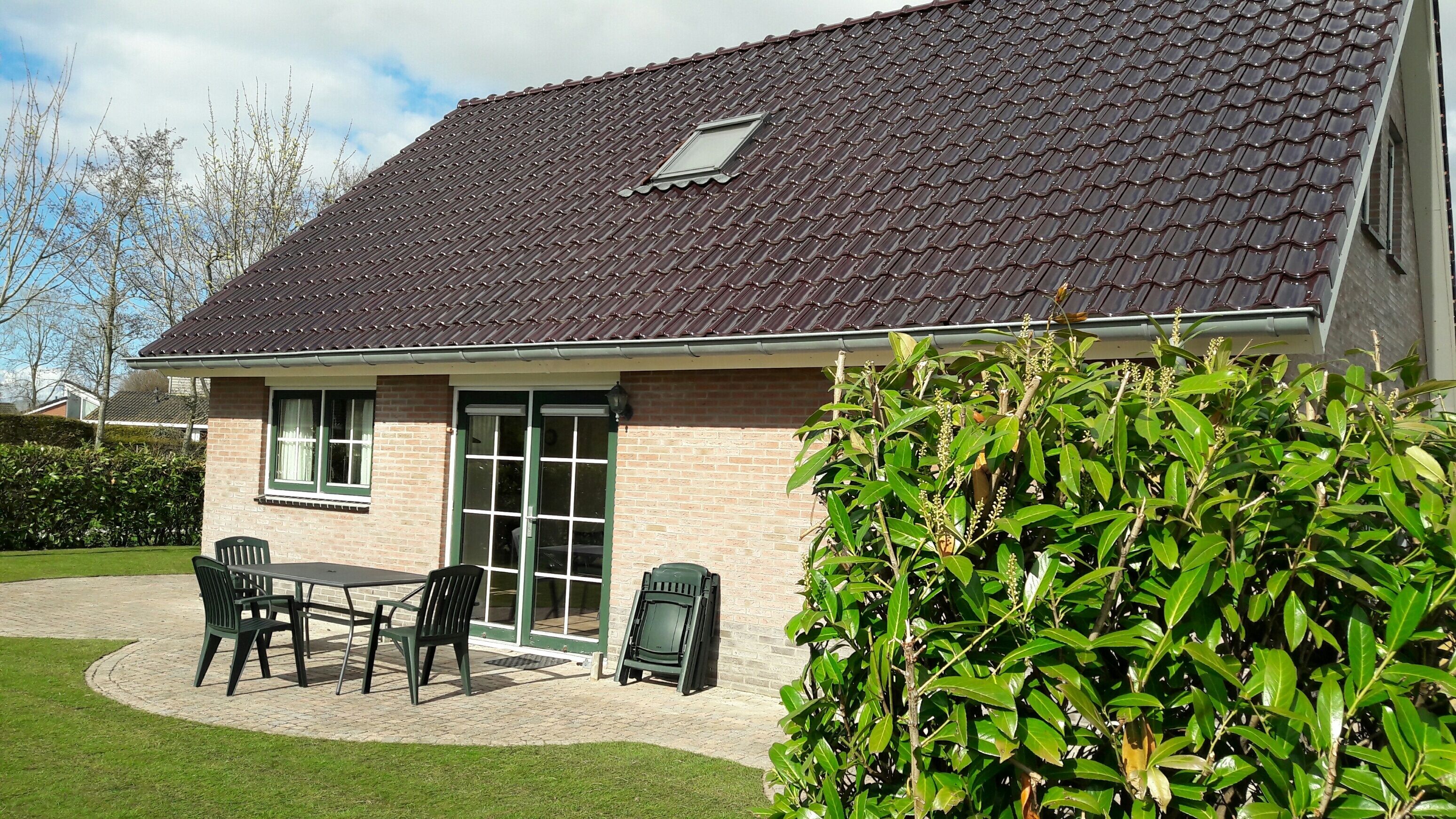 A house with a tiled roof, brick walls, and a small window. There is a patio with chairs and a table, and a green bush in the foreground.
