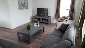 Ein Wohnzimmer mit grauem Sofa, einem dunkelholzigen Couchtisch, einem Flachbildfernseher auf einer grauen Unterhaltungsecke und einem Korbstuhl.