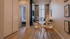 Een moderne eetruimte met een glazen tafel en witte stoelen, uitzicht op de stad door grote ramen, en een deur naar een andere kamer.