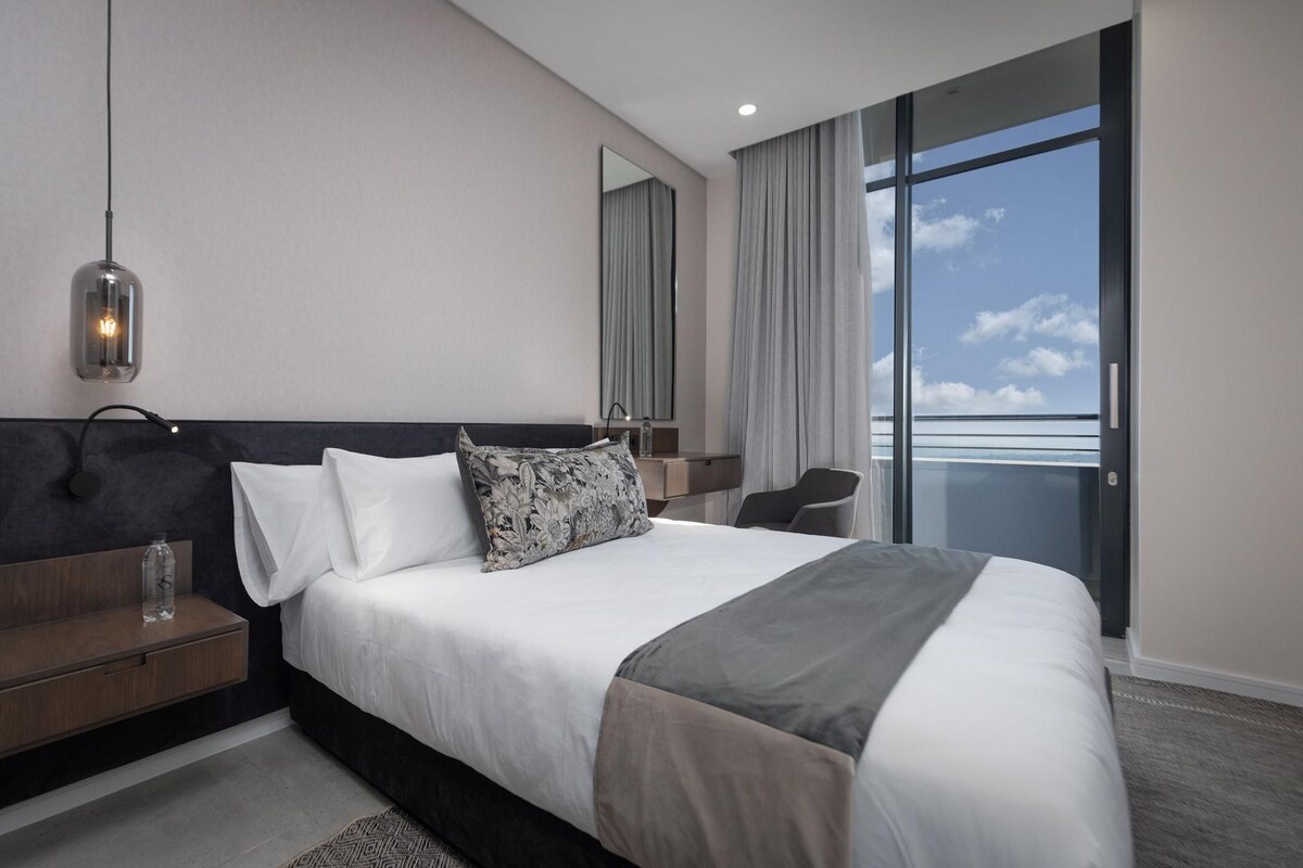 A modern hotel room with a large bed, a bedside table, a mirror, a window with a view, and a hanging light fixture.
