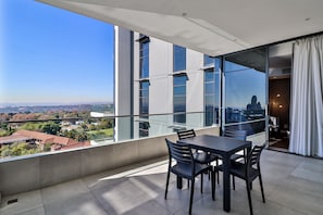 A balcony with a table and chairs, large windows offering a city view, and a glass door leading to an interior space. - Reserved Suites Illovo (Sandton)