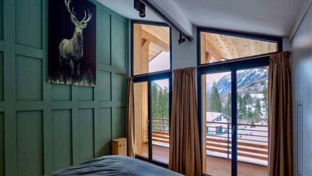 A bedroom with a large bed, a painting of a deer, and a view of a deck and mountains through large windows. - Chalet nestled above the slopes at Les Houches (Les Houches)