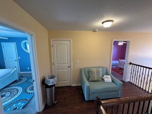 A room with a blue accent wall, a white door, a teal armchair with a striped pillow, and a trash can. - A family home that is perfect for any trip to beautiful Savannah! (Savannah)