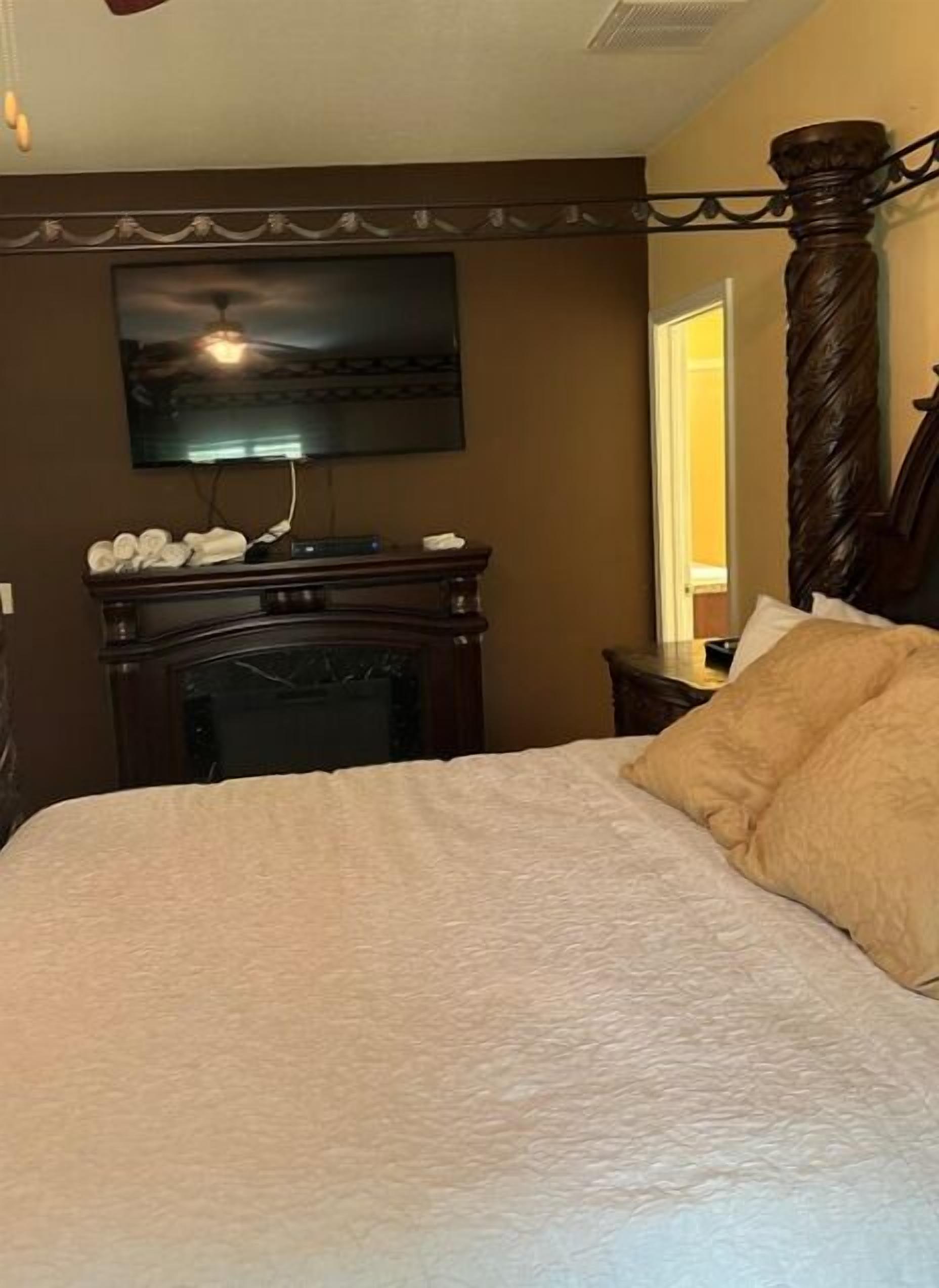 A bedroom with a large bed, a fireplace, a television mounted on the wall, and a door leading to another room.