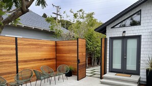A modern outdoor seating area with metal chairs and a glass table, adjacent to a building with a grey facade and a large glass door. - Beautiful 2 Bedroom Home in Santa Monica (Santa Monica)