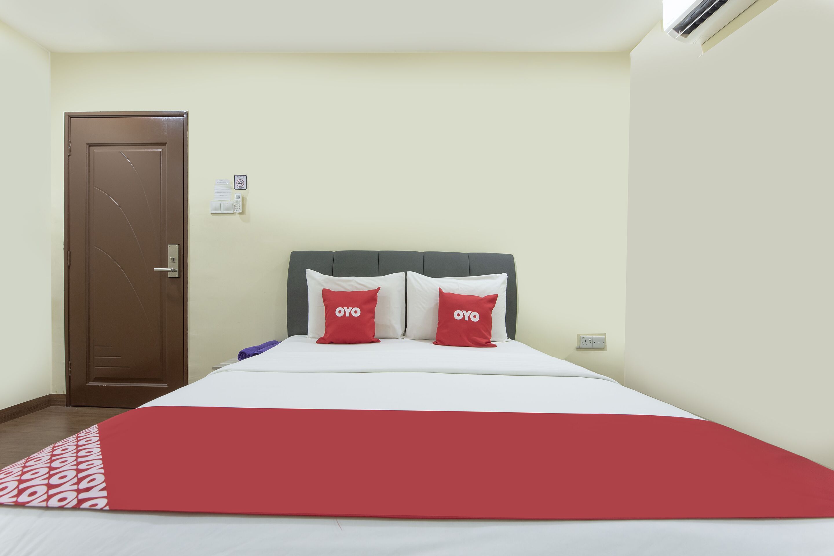 A hotel room with a bed, a brown door, a wall-mounted thermostat, and a light switch.