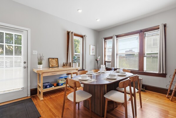 A dining area with a wooden table set for four, surrounded by chairs, with a window on one side and a door on the other.