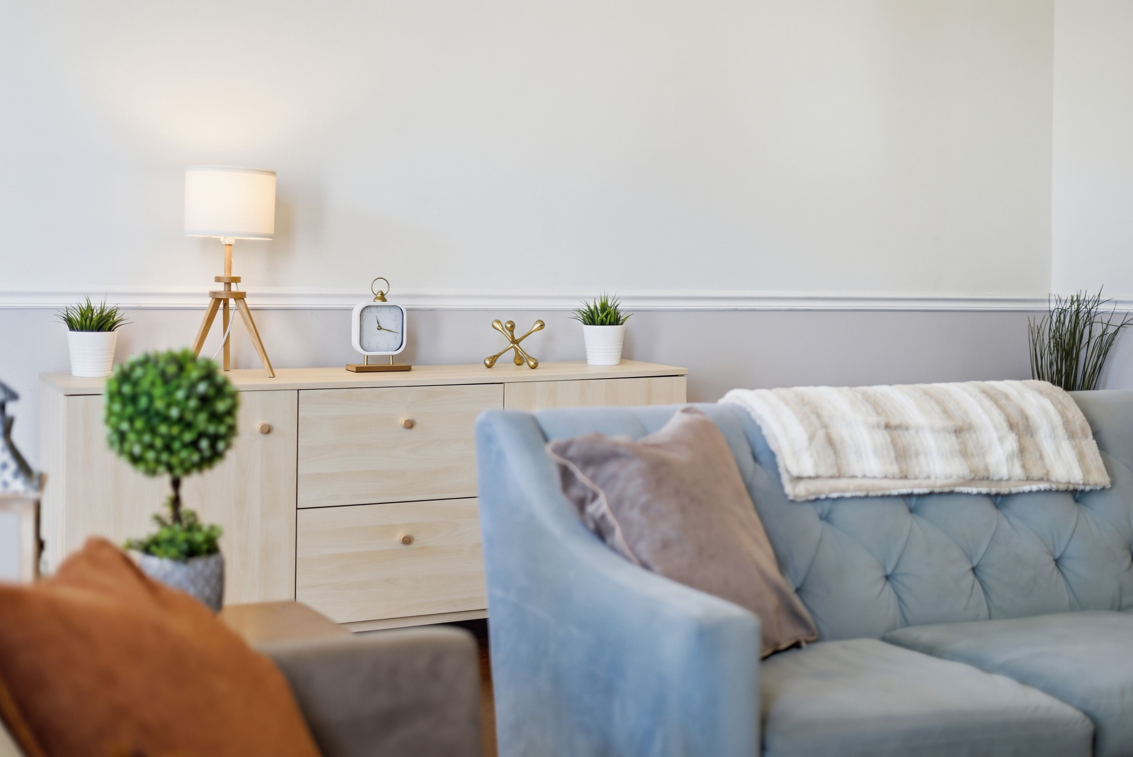 A living room with a light blue sofa, a small round table with a lamp, and a wooden dresser with two potted plants.