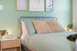 Uma cama bem arrumada com estrutura de metal, uma mesa de cabeceira com abajur e uma parede com quadros.