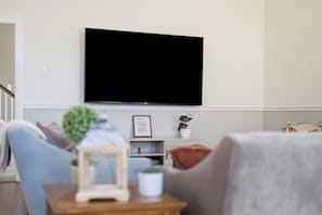 A living room with a flat-screen TV mounted on the wall, a small wooden side table with a picture frame and a plant, and a grey armchair.