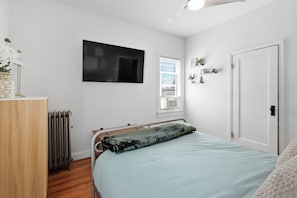 A bedroom with a bed, a radiator, a TV, a window with blinds, and a door.