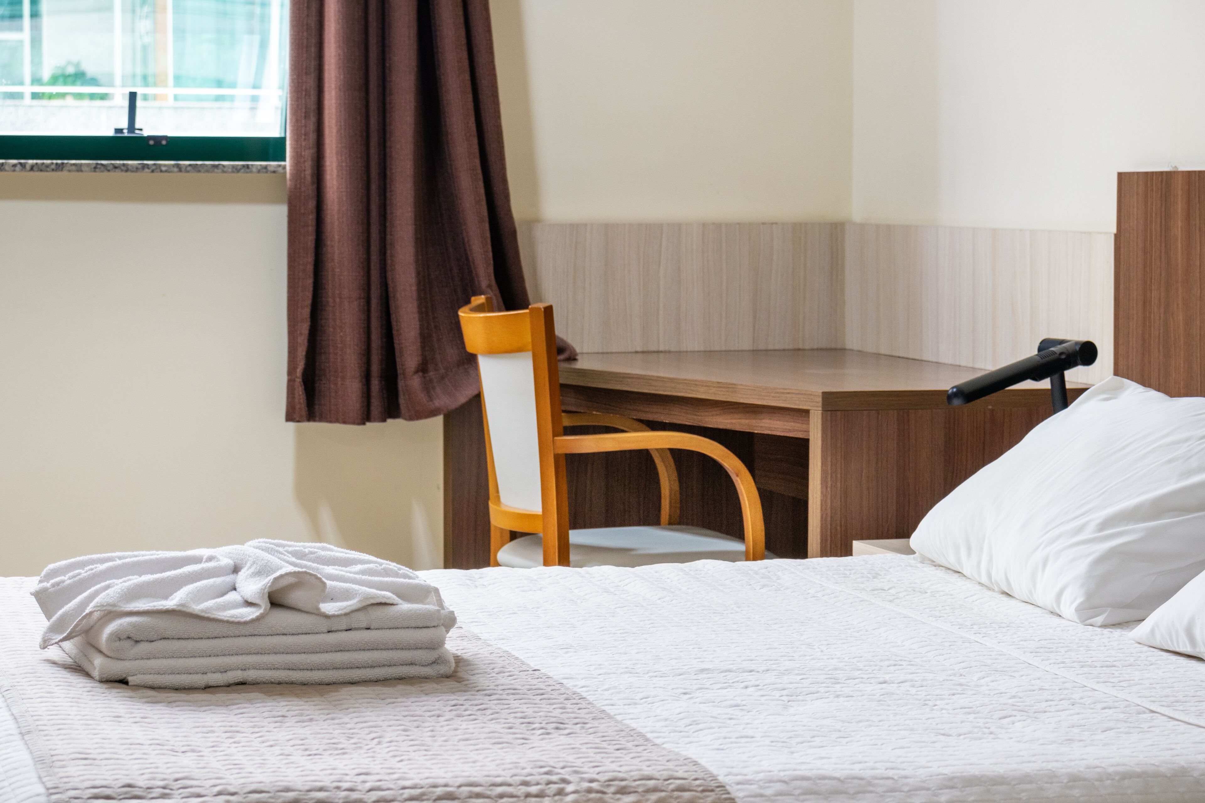 A neatly made bed with a folded white towel, a wooden desk, a chair, and a window with curtains.