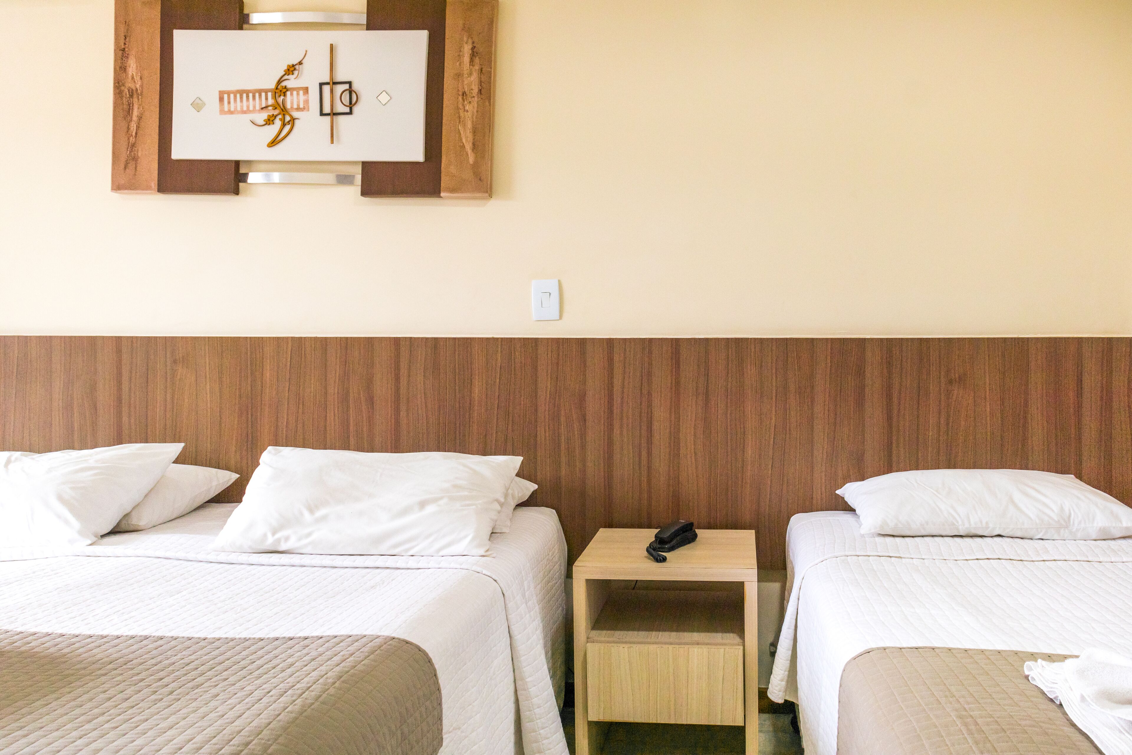 A hotel room with two beds, a wooden headboard, a bedside table with a phone, and a wall-mounted light switch.
