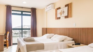 Habitación de hotel con dos camas, un escritorio y una silla. Se ve una ventana con cortinas, un equipo de aire acondicionado y un cuadro en la pared.