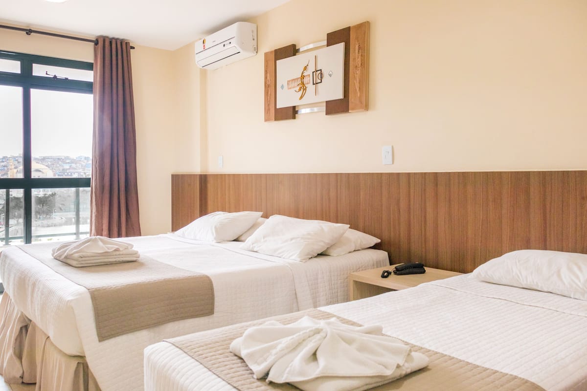 Habitación de hotel con dos camas, cabecero de madera, aire acondicionado de pared y una ventana con cortinas.
