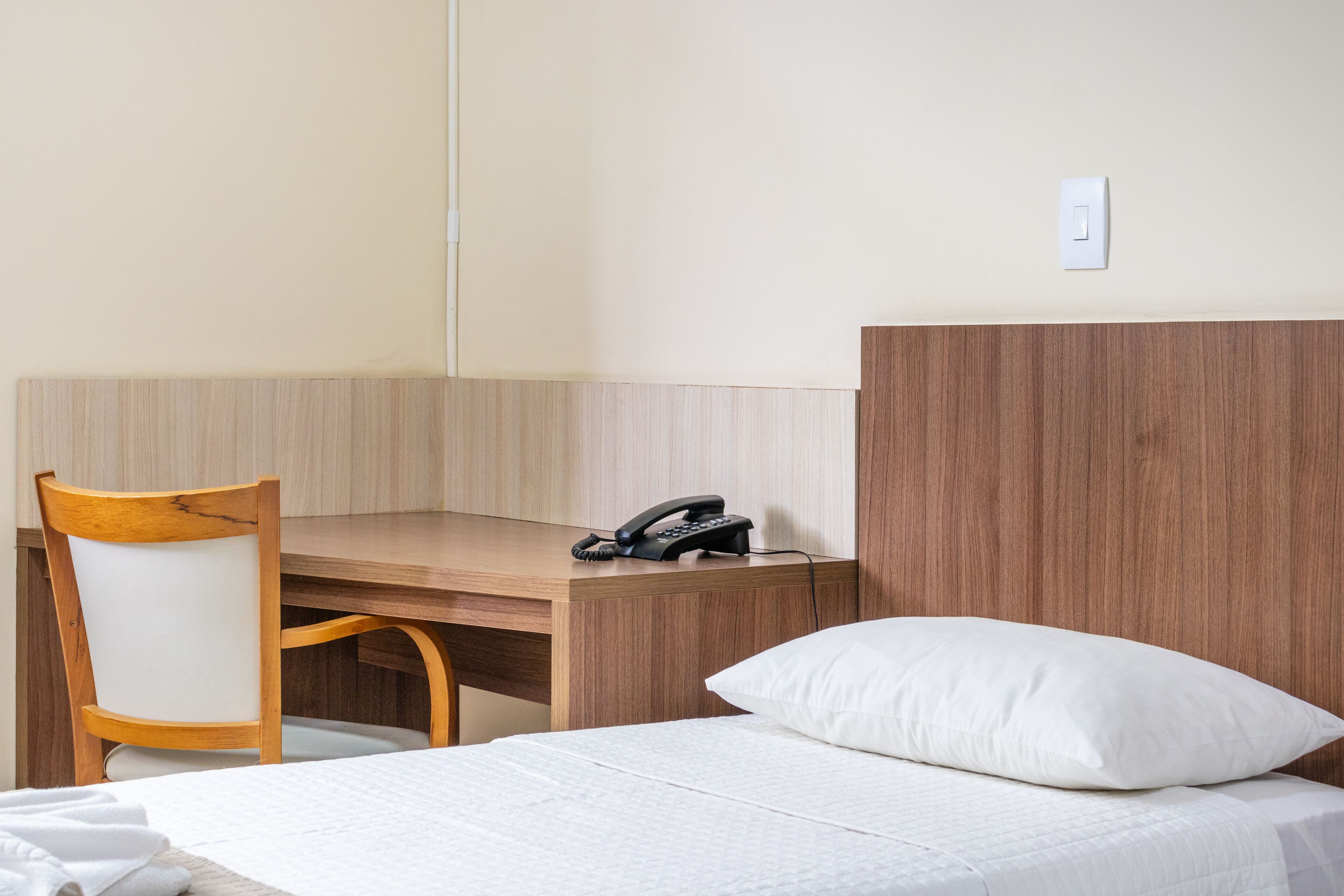 Quarto de hotel com escrivaninha de madeira, uma cadeira, uma cama com travesseiro branco e um telefone na escrivaninha.