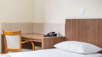 Quarto de hotel com escrivaninha de madeira, uma cadeira, uma cama com travesseiro branco e um telefone na escrivaninha.