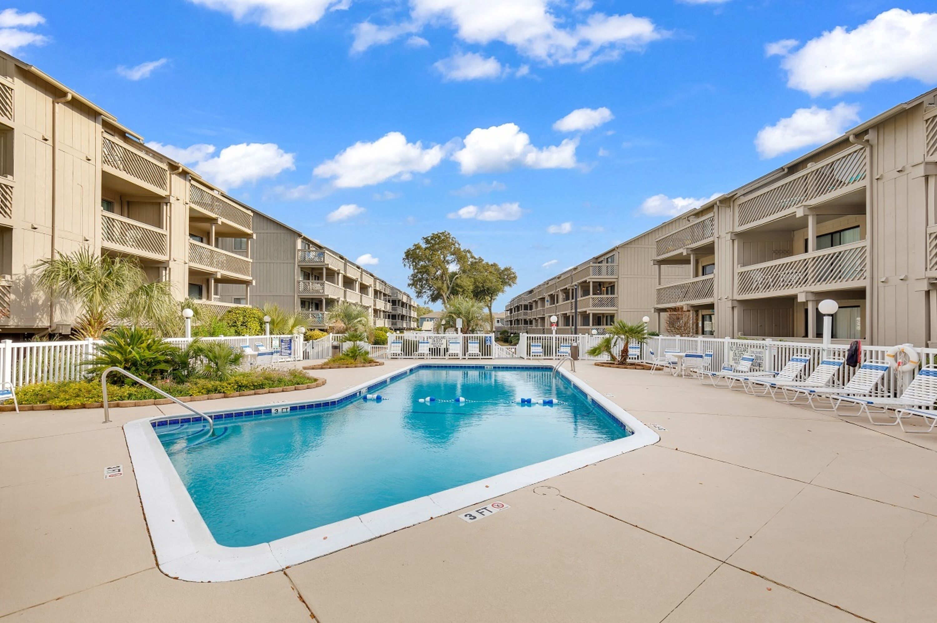 A swimming pool surrounded by a building with balconies, palm trees, and lounge chairs.