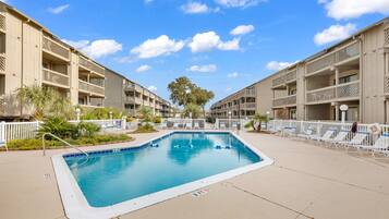 A swimming pool surrounded by a building with balconies, palm trees, and lounge chairs.