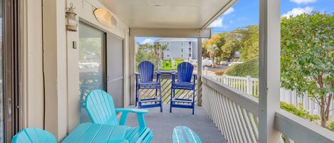A covered balcony with turquoise chairs and a table, overlooking a residential area with a white fence and greenery.