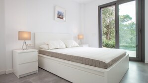 Un dormitorio moderno con una cama grande, mesitas de noche blancas y vistas a la vegetación a través de un ventanal corredero.