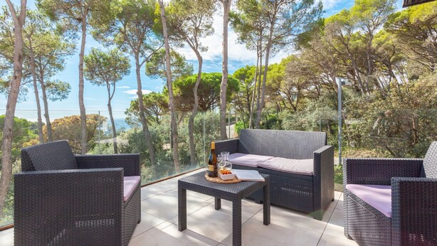 Una terraza con muebles de mimbre, una mesa con botella y vaso, y vistas a árboles y al mar.