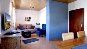 Un salon comprenant un canapé, un fauteuil bleu, une table à manger en bois et un téléviseur fixé au mur.
