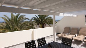 A covered outdoor seating area with lounge chairs and a table, overlooking a beach with palm trees and the sea. - Newly renovated penthouse in a complex with pool on the seafront (Costa Teguise)