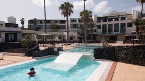 A swimming pool with a water slide, surrounded by palm trees and a stone wall. - Newly renovated penthouse in a complex with pool on the seafront (Costa Teguise)