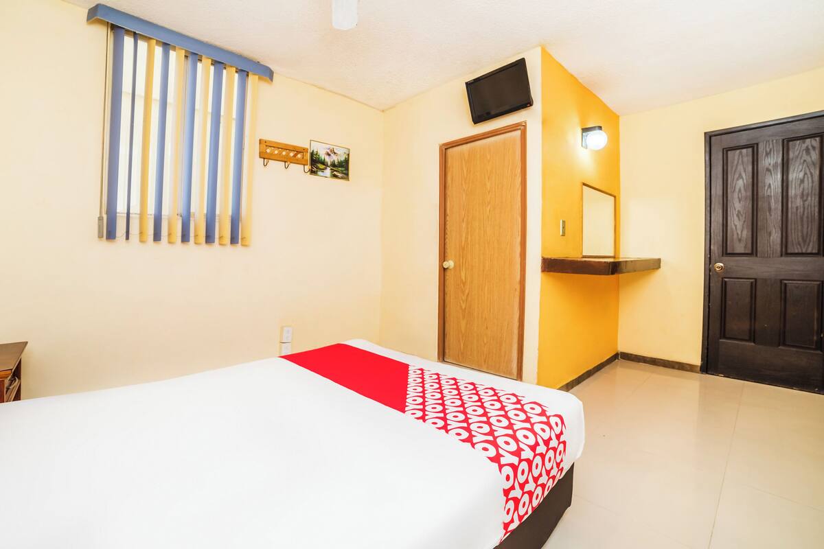 Habitación de hotel con una cama, una puerta de madera, un televisor montado en la pared y una ventana con persianas.