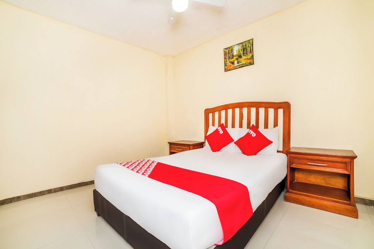 Una cama bien hecha con una colcha roja y blanca, dos mesitas de noche de madera y un cuadro en la pared.